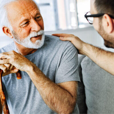 Happy mature man sitting, holding cane, speaking with younger man just off camera.