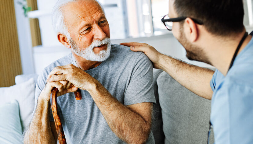 Happy mature man sitting, holding cane, speaking with younger man just off camera.