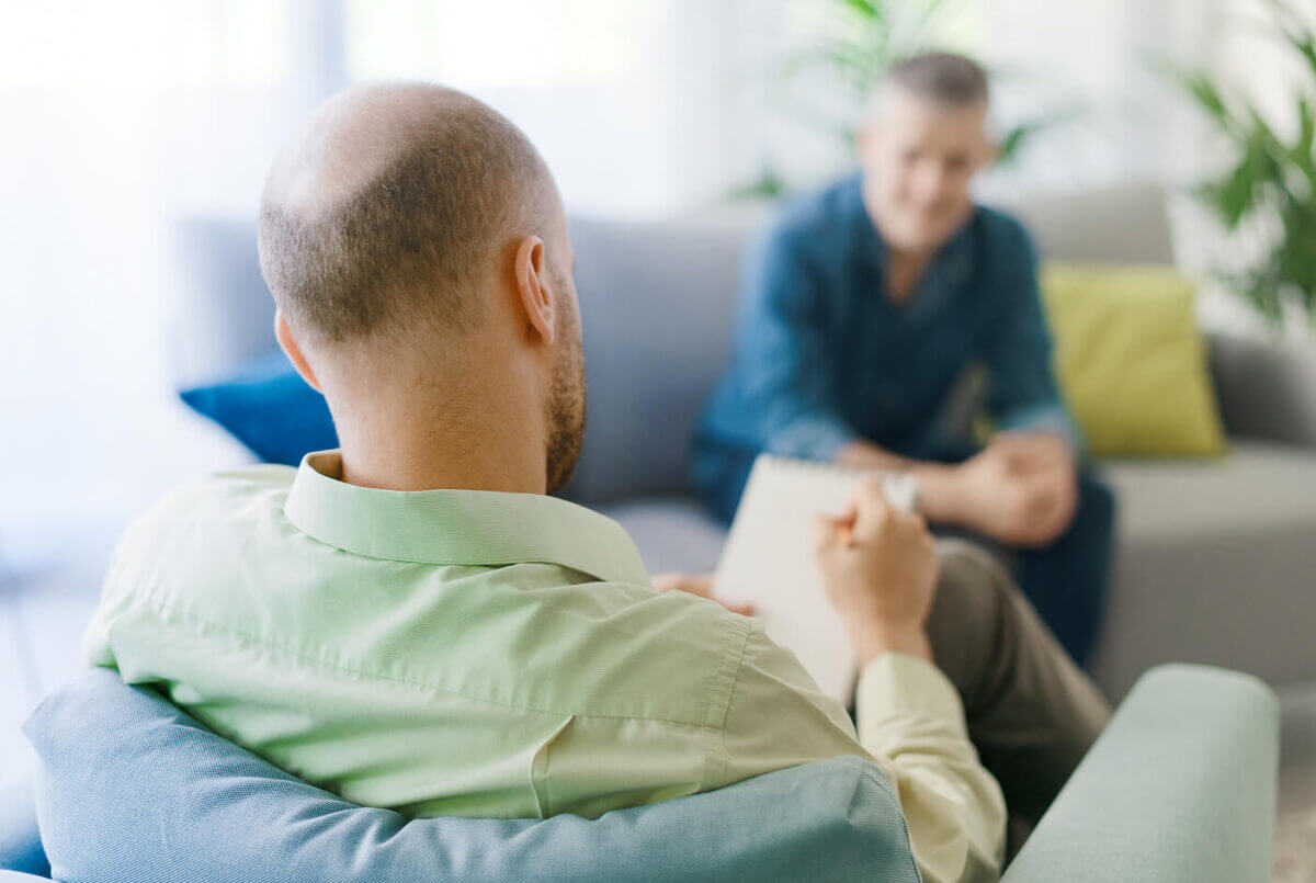 Mature man, out of focus in background, in foreground camera is looking over the shoulder of a younger man writing on a notepade while engaged with the mature man.